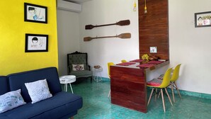 Una habitación con un sofá azul, una mesa de comedor de madera con sillas, una mesita redonda y dos cuadros en una pared amarilla.