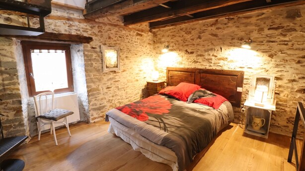 A rustic bedroom with a stone wall, wooden ceiling beams, a bed with a floral bedspread, a chair, and a bedside table with a lamp.