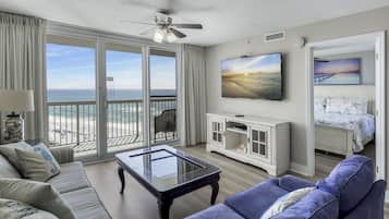 A spacious living room with a large glass coffee table, a sofa, and a television mounted on the wall. There is a balcony with a view of the beach and ocean.