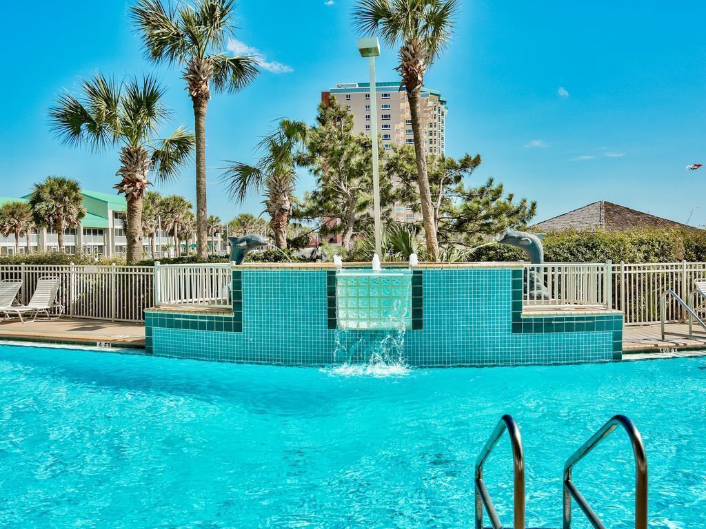 A swimming pool with a water fountain, surrounded by palm trees and a tall building in the background.