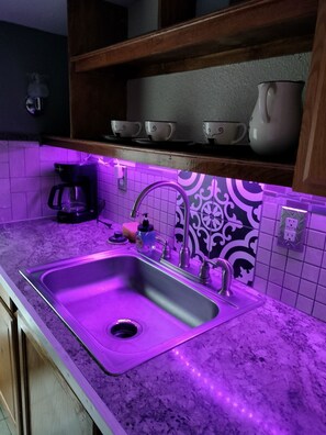 A kitchen with a stainless steel sink, purple under-cabinet lighting, a coffee maker, and decorative tiles. - Experience comfort near sports action, 4BR/1Bath home by Arrowhead 14 min. (Independence)