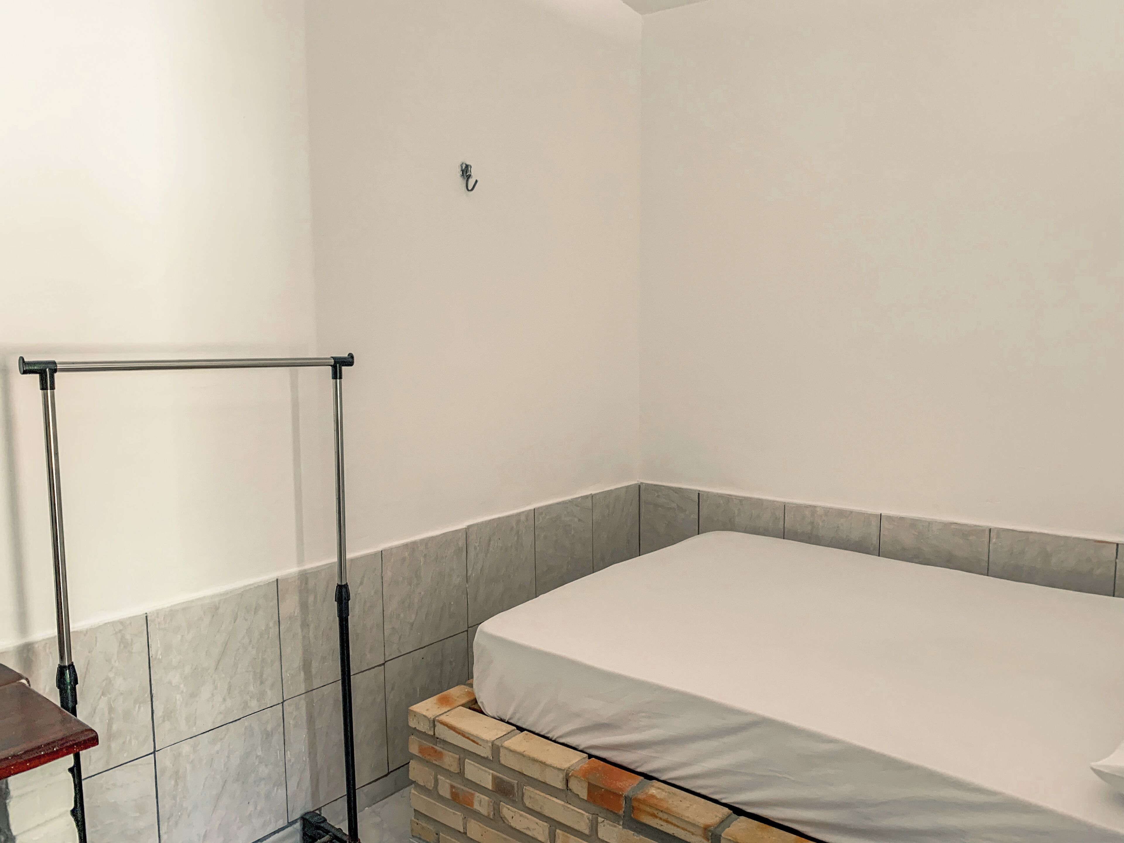 Una habitación pequeña con una cama individual, un perchero metálico en la pared, un banco de madera y una sección inferior revestida con baldosas.