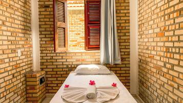 Una habitación pequeña y acogedora con pared de ladrillo, una cama individual con ropa blanca y una ventana con persianas rojas.