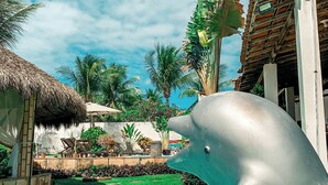 A dolphin statue in a swimming pool with a tropical garden and a thatched-roof structure in the background. - Pousada La Goduria (Aracati)