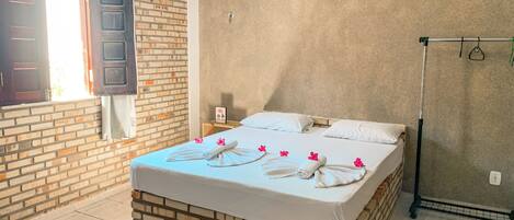 Habitación de hotel con pared de ladrillo decorativa, cama con ropa blanca y flores rosadas, un perchero y una ventana con la puerta entreabierta.