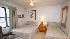 Camera da letto con due letti, una sedia, un comodino, una lampada, un poster appeso al muro e una finestra con tende.