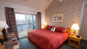 A bedroom with a large bed, two bedside lamps, a wooden nightstand, a quilted bench, and a view of a deck through a glass door.