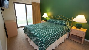 A bedroom with a green bedspread, two bedside lamps, a television on the wall, and a sliding glass door leading to a balcony. - Ski-In/Ski-Out Condo with Access to Indoor Pool (Ludlow)