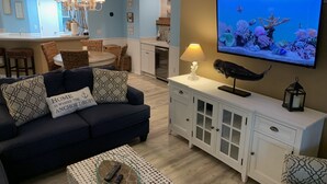 Una sala de estar con un sofá azul oscuro, un televisor que muestra una escena submarina, una lámpara y un pájaro en un pedestal.