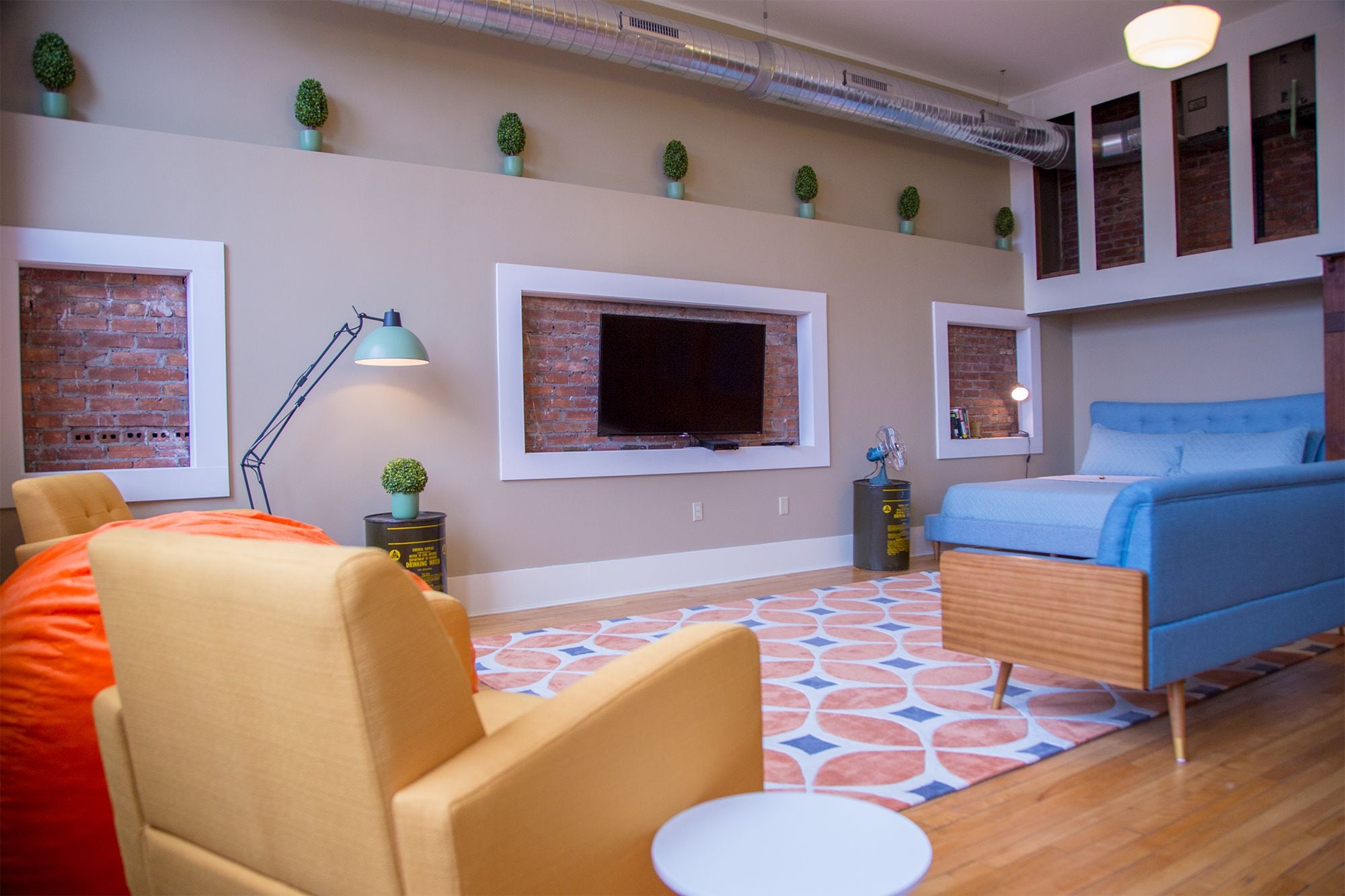 A modern bedroom with a bed, a desk, and a chair. There are multiple windows and a brick wall.