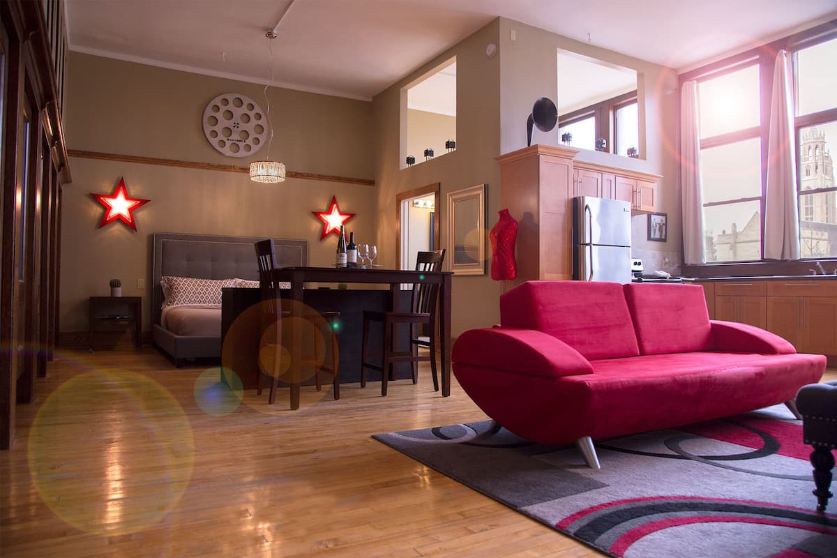 A modern living room with a red sofa, a dining area with a table and chairs, a kitchen with a refrigerator, and a large window.