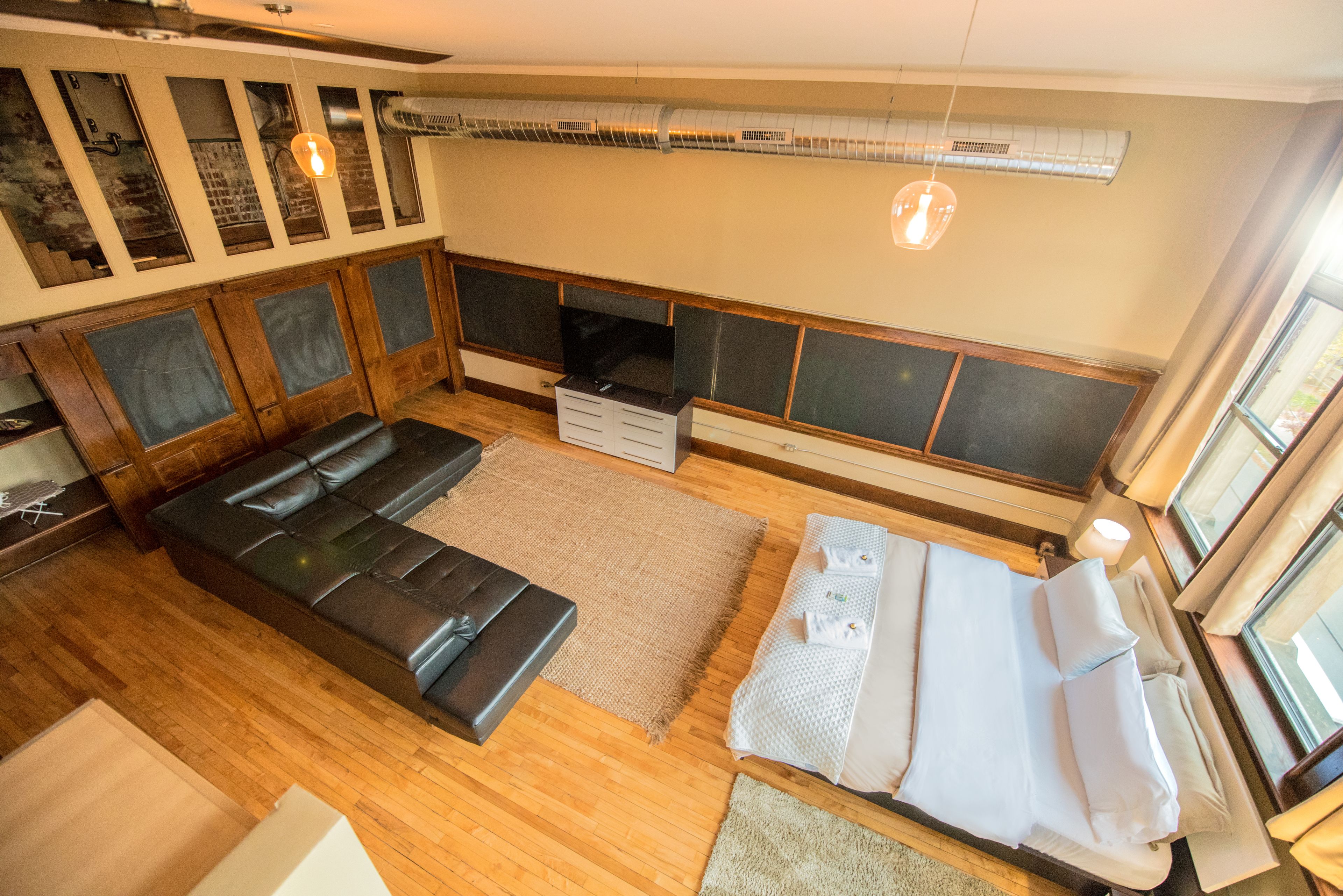 A room with a black leather sofa, a bed with white linens, a wooden floor, and a large window.
