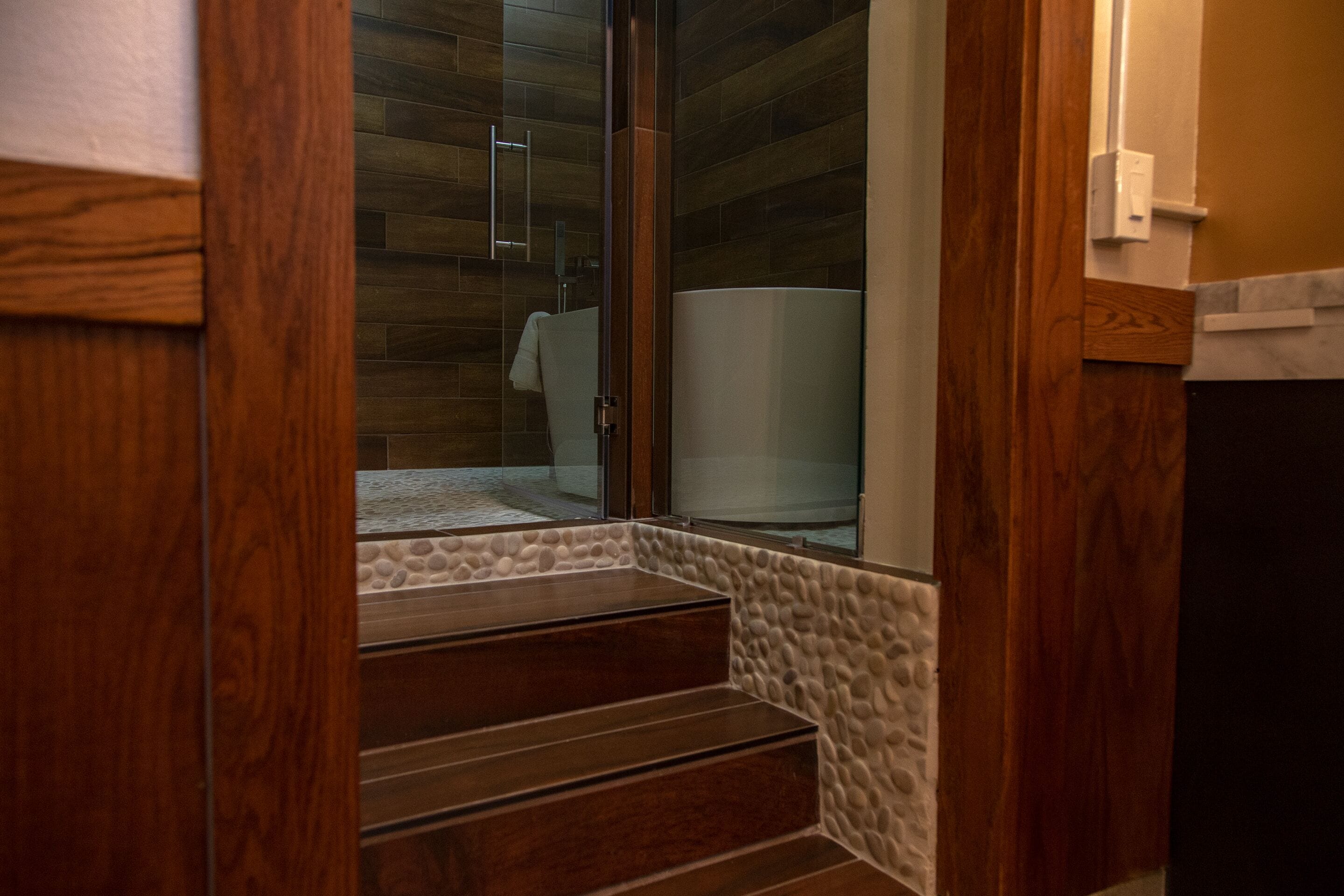 Un escalier en bois mène à une salle de bain dont un des murs est en pierre et qui est équipée d’une douche à parois de verre.