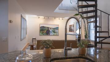 A kitchen with granite countertops, a stainless steel sink, and a wooden bench. There is a painting on the wall and a spiral staircase to the right.