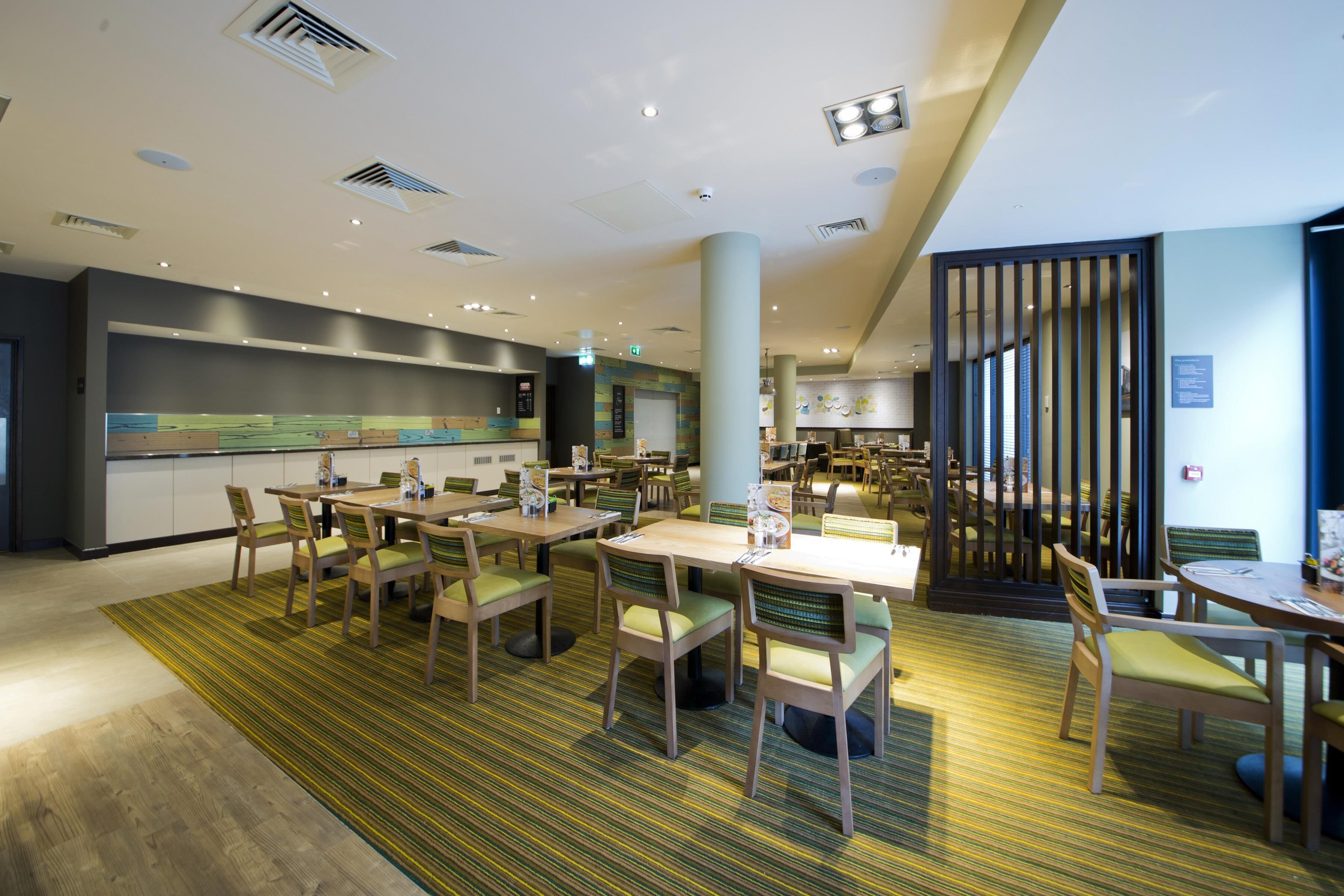 A modern hotel lobby with multiple dining tables and chairs, a large pillar, and a view of the kitchen area.