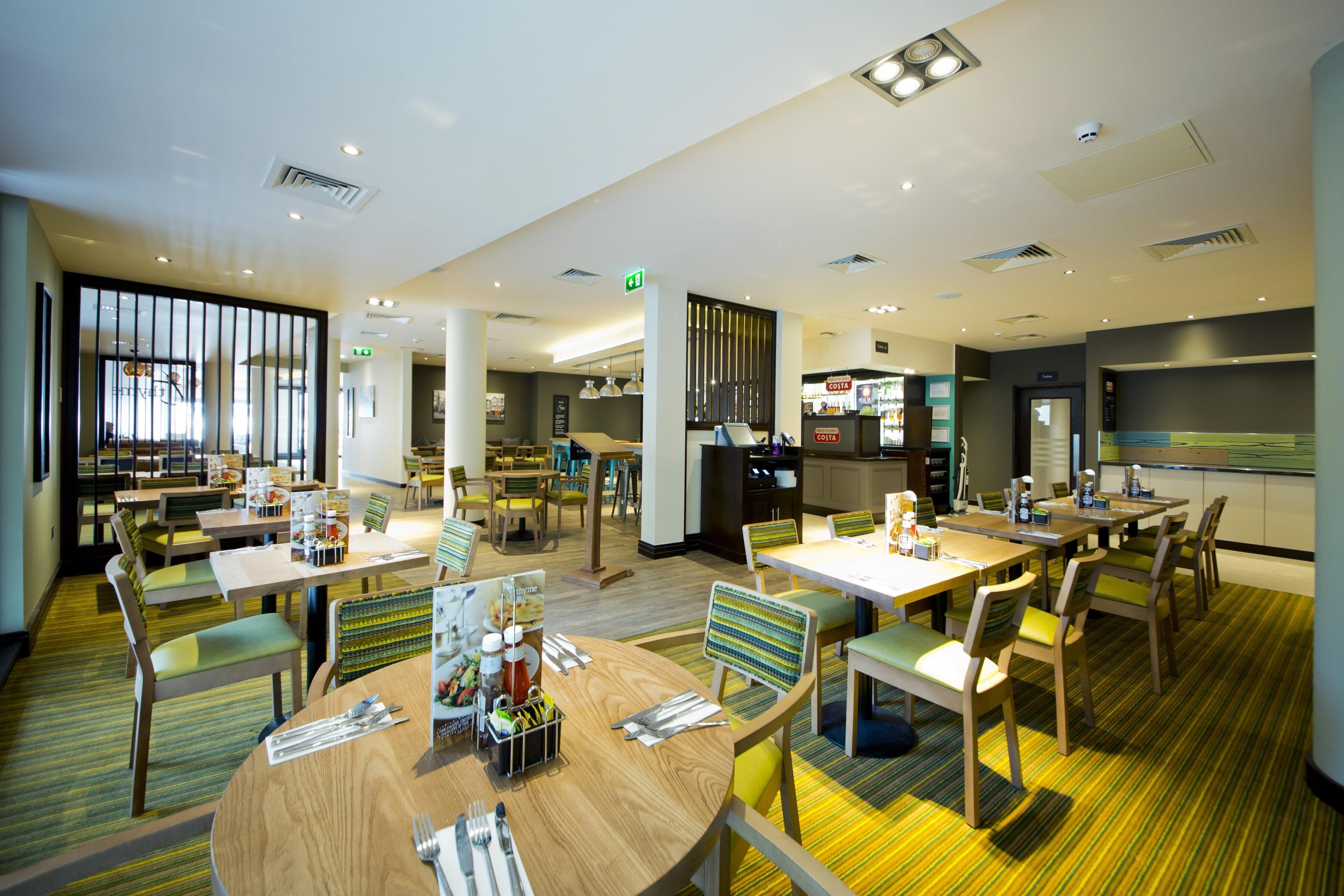 A modern hotel dining area with multiple tables, chairs, and a kitchen in the background.