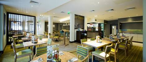 A modern hotel dining area with multiple tables, chairs, and a kitchen in the background.