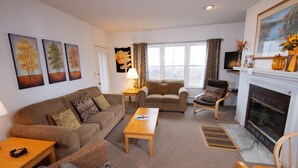A living room with a sofa, armchair, coffee table, and fireplace. There are three paintings on the wall, a lamp, and a television. - Okemo Retreat! Ski-In/Ski-Out Condo (Ludlow)