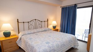 A neatly made bed with a floral comforter, two bedside lamps, a wooden nightstand, and a view through a large window with blue curtains. - Okemo Retreat! Ski-In/Ski-Out Condo (Ludlow)