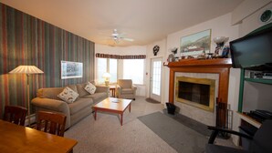 A living room with a sofa, armchair, coffee table, and fireplace. There is a television on a stand, a lamp, and a painting on the wall. - Mountainside Suite! Arcade, Pool, Hot Tub Access (Ludlow)