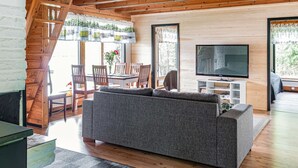 Un salon moderne avec un canapé gris, des poutres en bois au plafond, un téléviseur et un coin repas comprenant une table et des chaises.