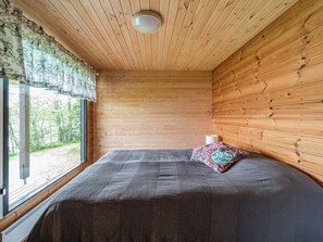 A room with wooden walls, a bed with a grey bedspread, a window with a view of greenery, and a white lamp on the nightstand. - Kotiranta by Interhome (Petäjävesi)