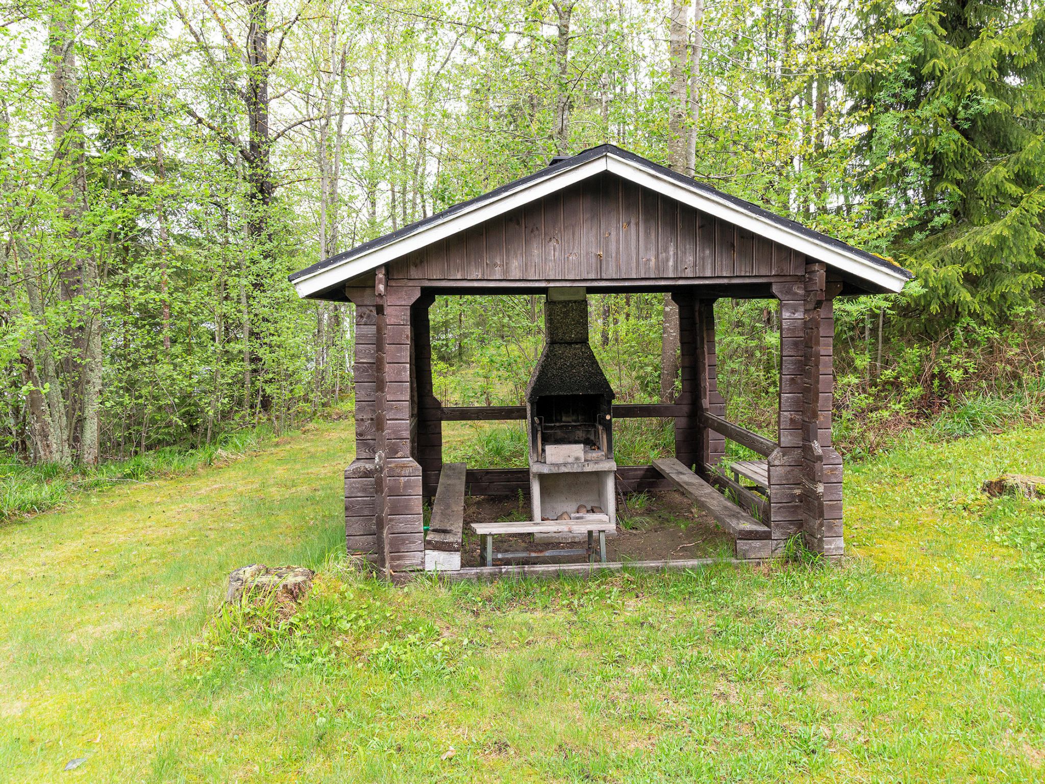 A rustic wooden gazebo with a fireplace, surrounded by a forested area.