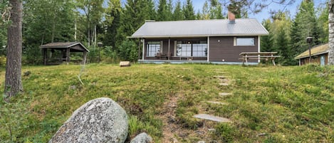 Une cabane avec une véranda, une table de pique-nique et un petit abri de jardin, située dans une zone boisée.