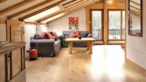 A cozy loft-style living room with a wooden ceiling, a sofa set, a coffee table, and a large window offering a view of the outdoors.