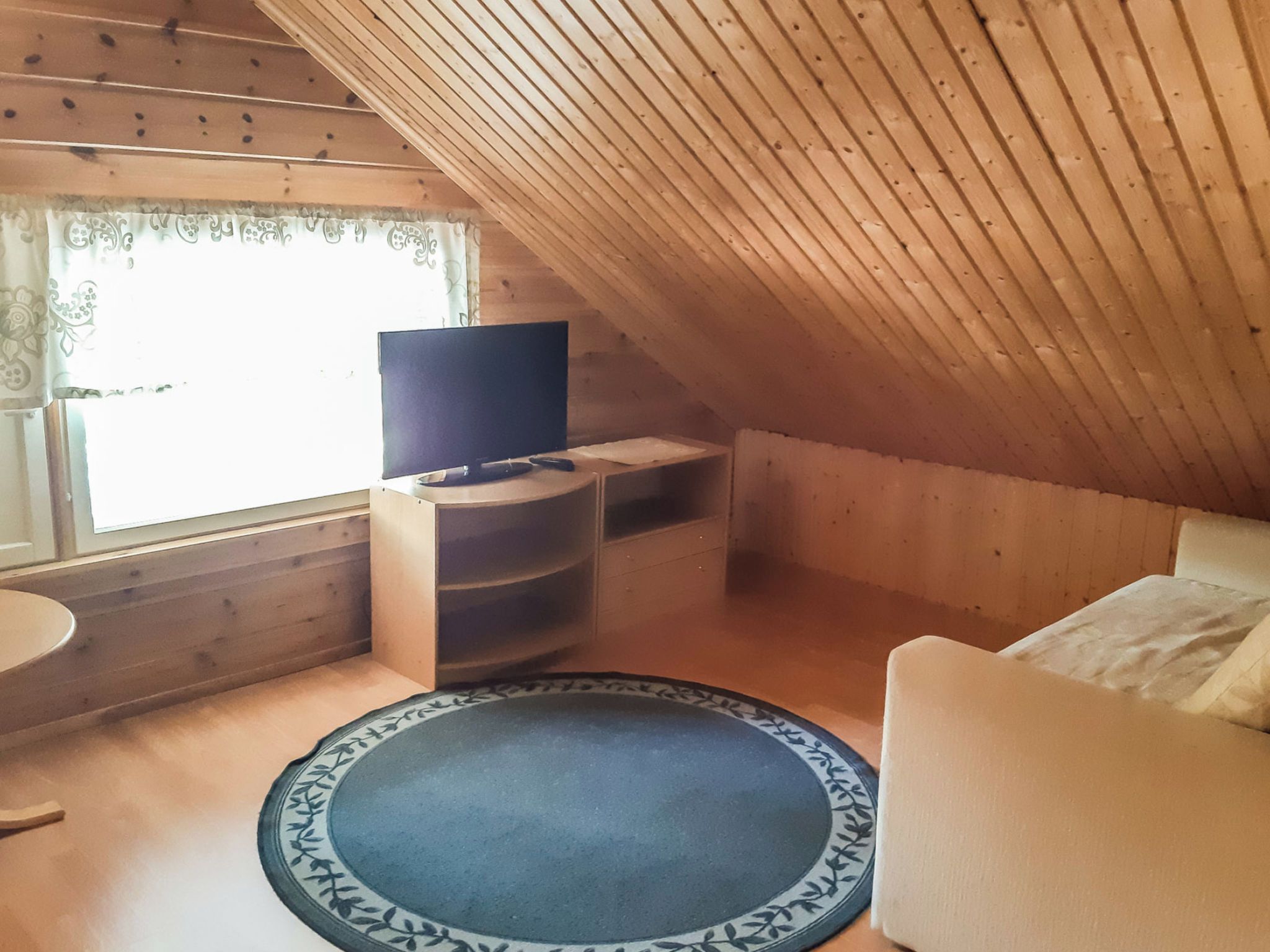Pieni, viihtyisä ullakkohuone, jossa on televisio puisen jalustan päällä, pyöreä matto ja valkoinen sohva.