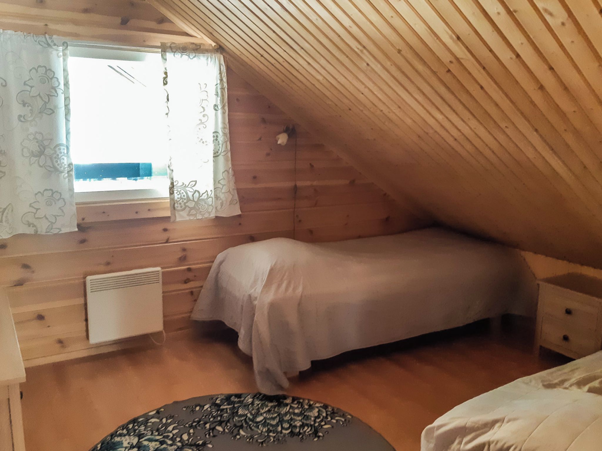 A small, cozy attic bedroom with a sloped ceiling, a single bed, a radiator, and a window with curtains.