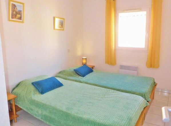 A room with two single beds, each with a blue pillow, a bedside table, and a window with yellow curtains. - Plein Sud by Interhome (Narbonne-Plage)