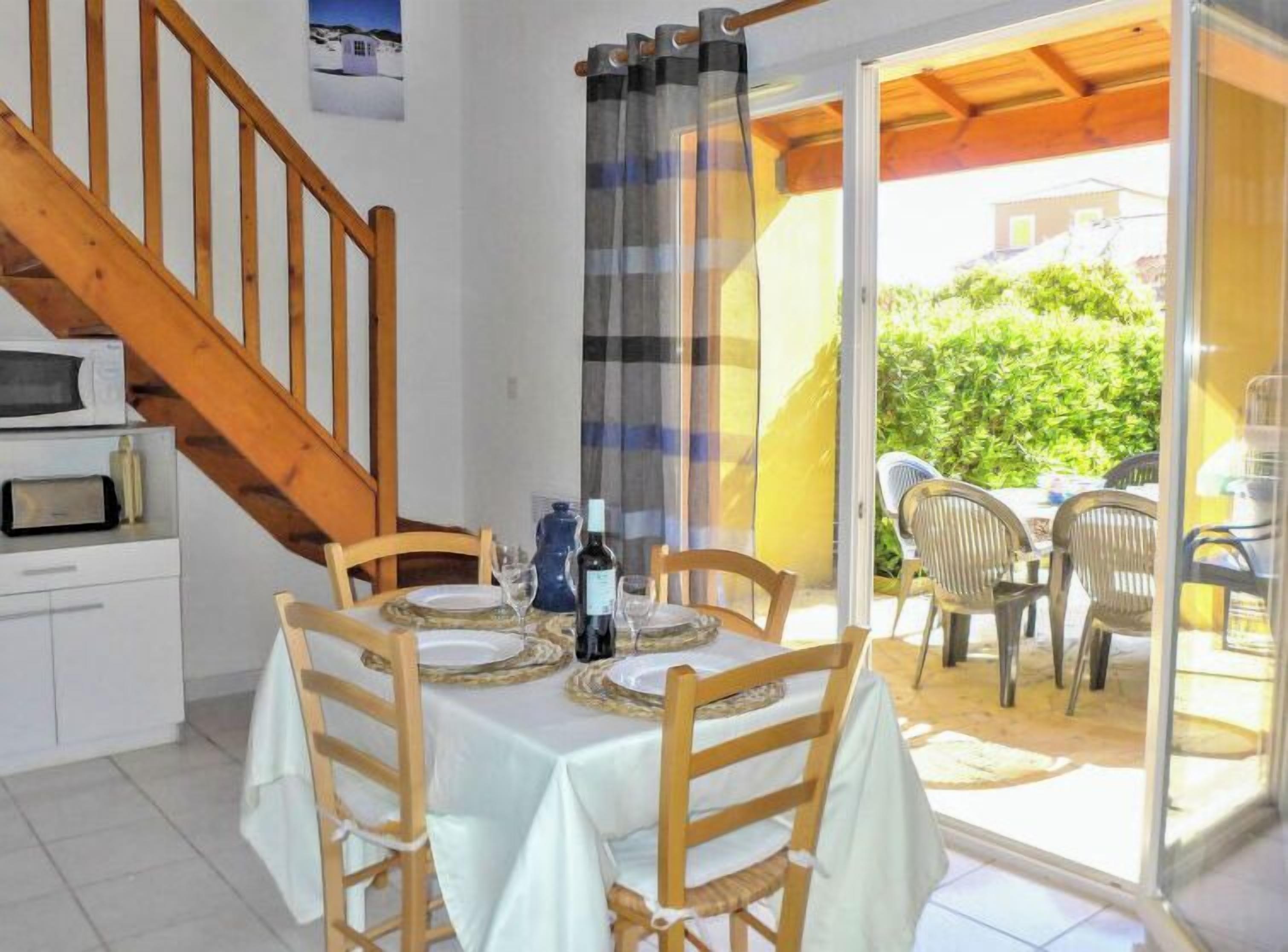 Un área de comedor con una mesa dispuesta para cuatro personas, sillas de madera, una botella de vino y una copa. Una escalera conduce a un nivel superior.