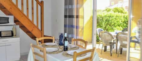 A dining area with a table set for four, wooden chairs, a bottle of wine, and a glass. A staircase leads to an upper level.