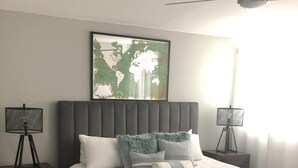 Ein ordentlich eingerichtetes Schlafzimmer mit einem großen Bett, einer Weltkarte als Wandbild darüber, einem Deckenventilator und zwei Stehlampen.