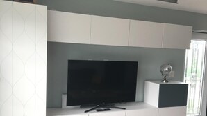 Ein modernes Wohnzimmer mit einem Flachbildfernseher auf einem weißen Schrank, Holzboden und einem Fenster mit Jalousien.