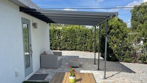 Eine Terrasse mit Holztisch und -bänken, ein überdachter Sitzbereich sowie ein weißes Gebäude mit Tür und Fenster.