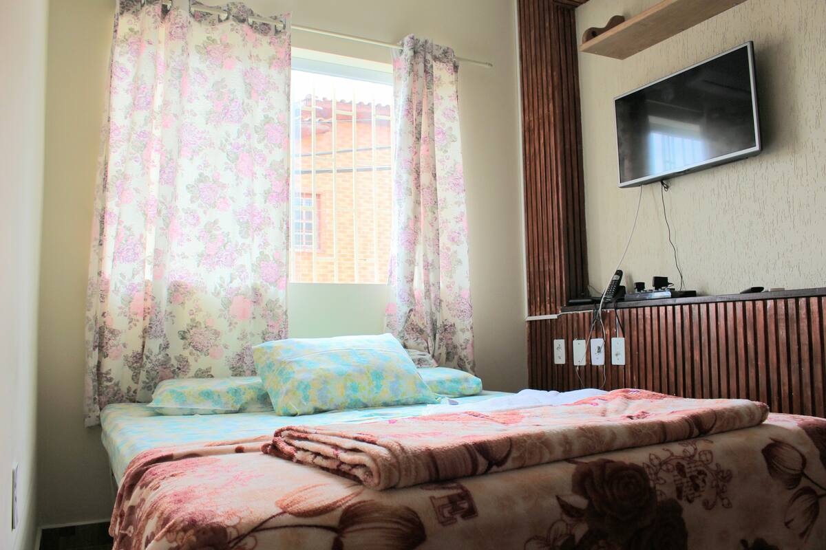 A bedroom with a bed, floral curtains, a television on the wall, and a window with a view of a brick building.