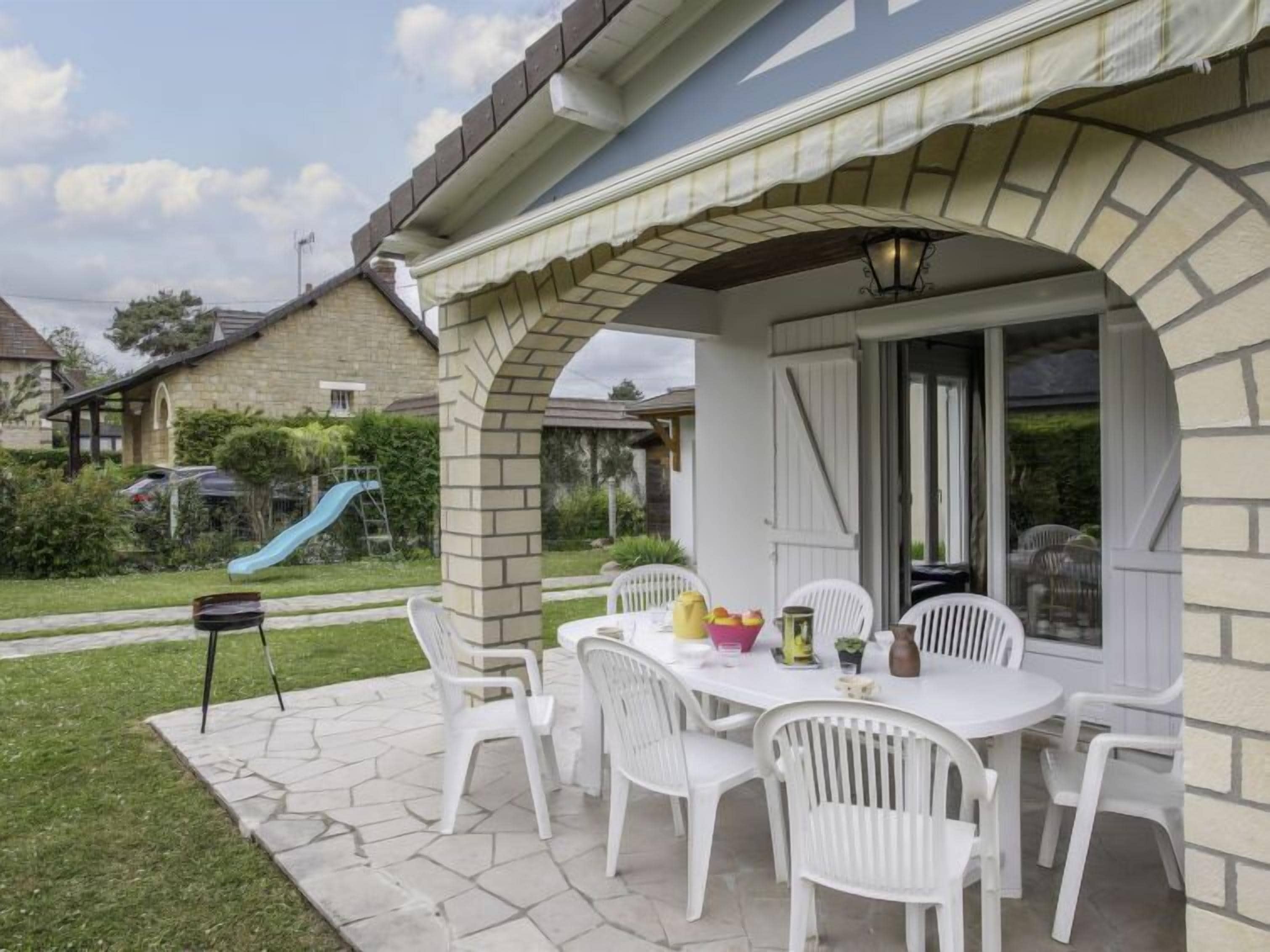 Ein Terrassenbereich im Freien mit Steinplattenboden, weißen Kunststoffstühlen und einem für das Essen gedeckten Tisch.
