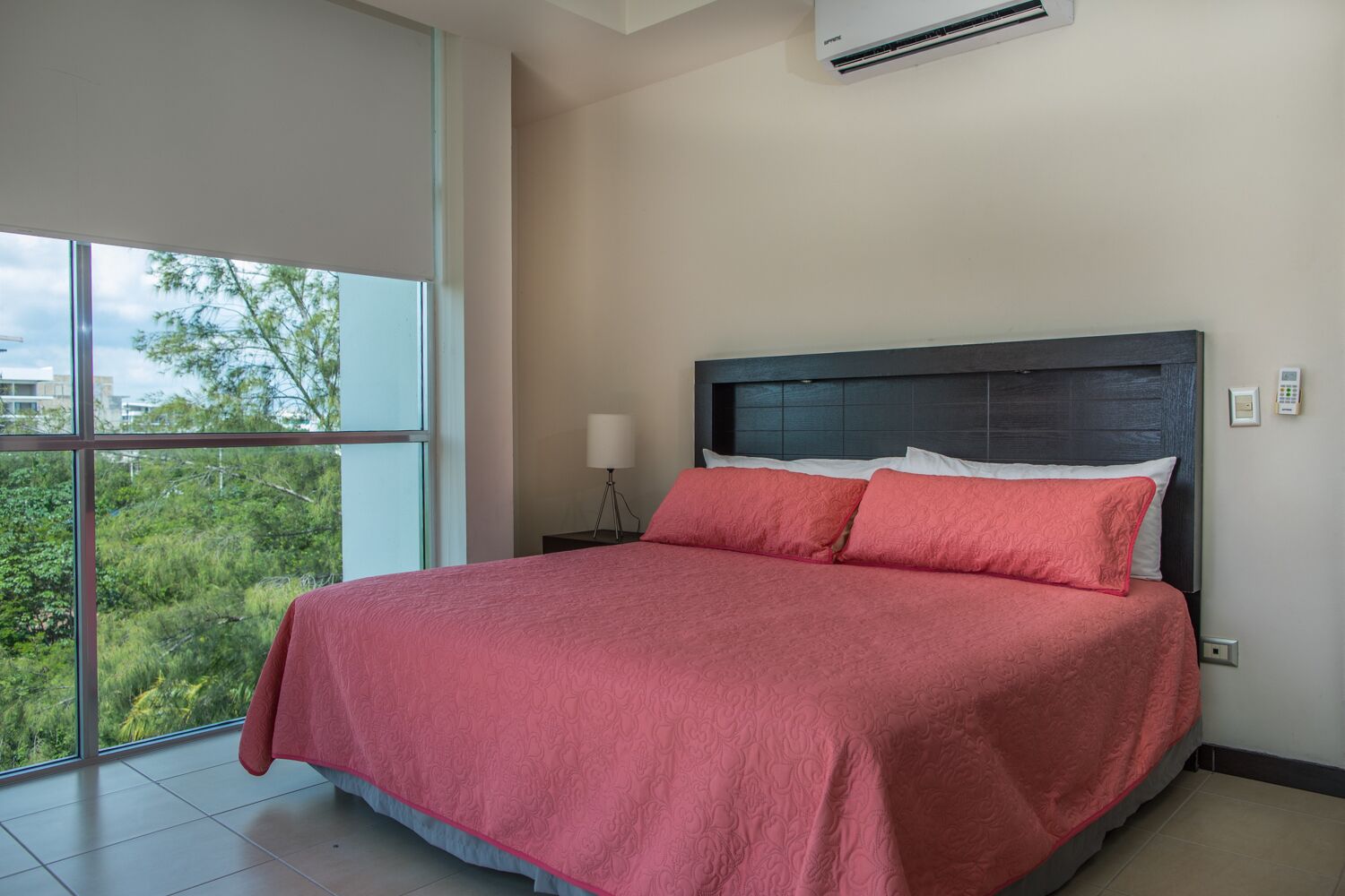 A modern bedroom with a large bed, a nightstand, a lamp, and a view of greenery through large windows.