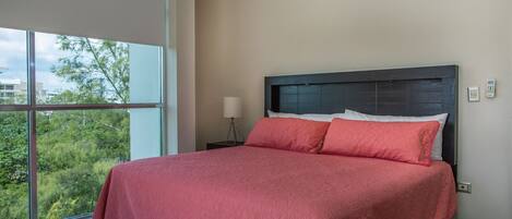 A modern bedroom with a large bed, a nightstand, a lamp, and a view of greenery through large windows.
