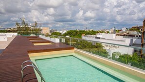 A rooftop swimming pool with wooden deck and glass railings, surrounded by buildings and greenery. - San Pedro Hotel 5ta Avenida (Playa del Carmen)