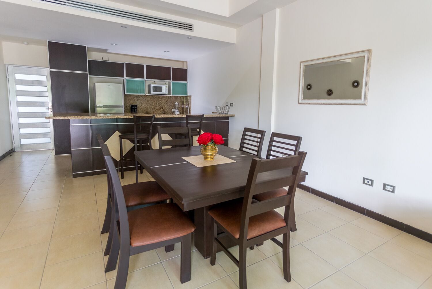 A modern dining area with a dark wooden table and chairs, a vase with red flowers, and a kitchen in the background.