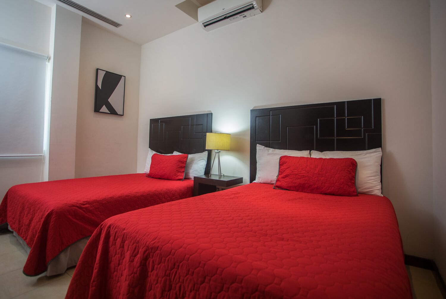 Habitación de hotel con dos camas, cada una con cubrecama rojo y almohadas a juego. Entre las camas, una mesita de noche con lámpara.
