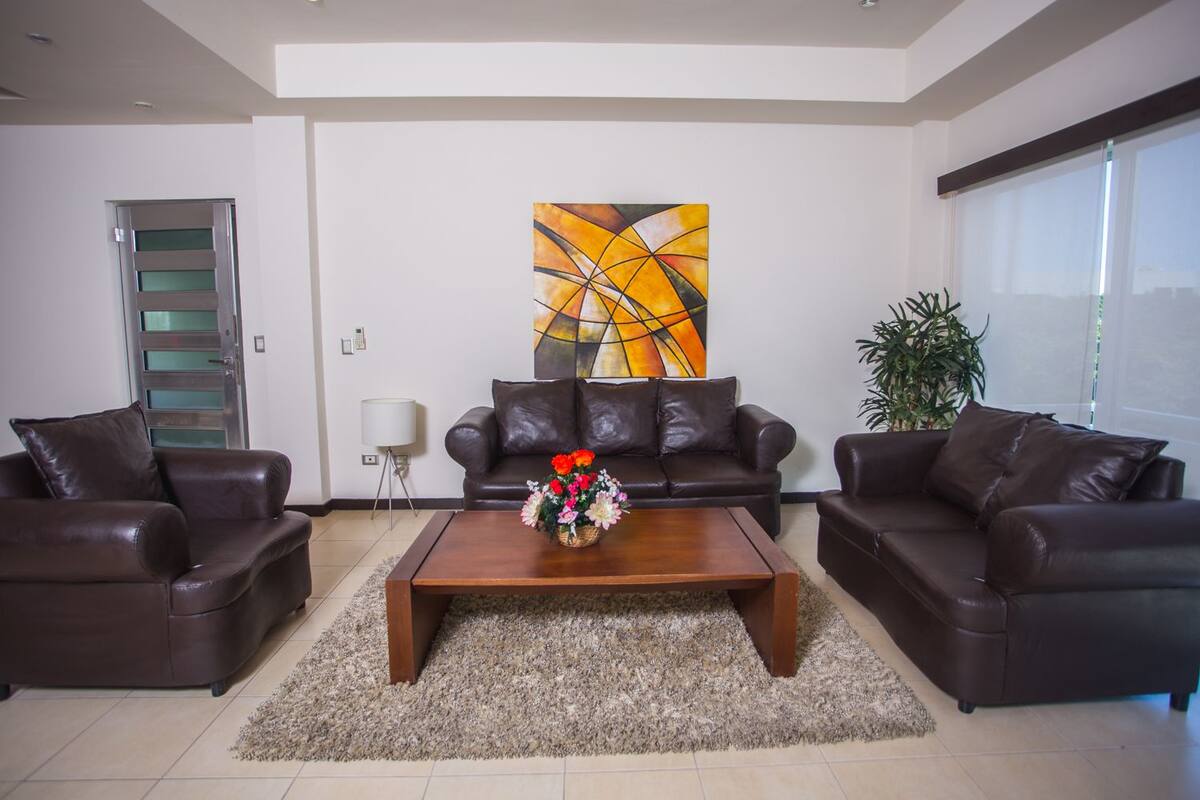 Ein modernes Wohnzimmer mit zwei Ledersofas, einem Holztisch mit floraler Dekoration, einem großen abstrakten Gemälde an der Wand und einer Zimmerpflanze.