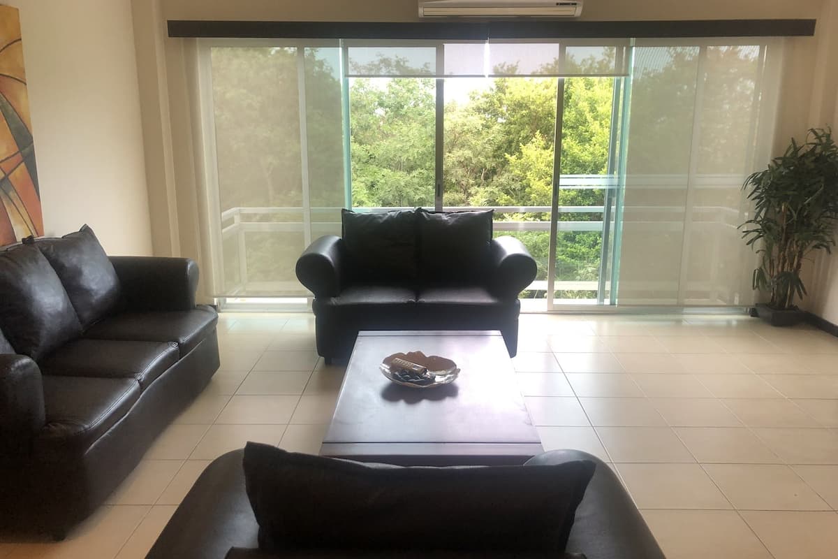 Ein geräumiger Wohnbereich mit zwei schwarzen Sofas, einem Couchtisch und einem großen Fenster mit Blick ins Grüne.