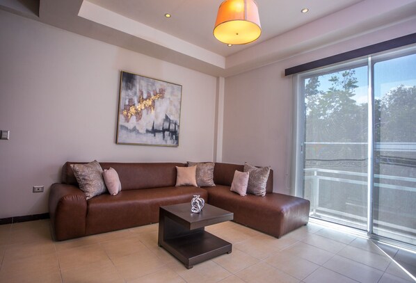 A modern living room with a brown leather sofa, a coffee table, and a large window offering a view of the outdoors.