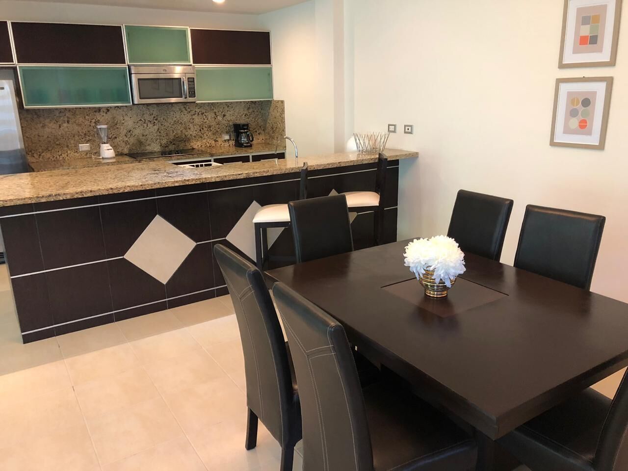 Una cocina moderna con encimera de granito, microondas integrado y cafetera. Hay una mesa de comedor con seis sillas y un jarrón con flores blancas. La cocina tiene un salpicadero negro y verde, y hay cuadros en la pared.