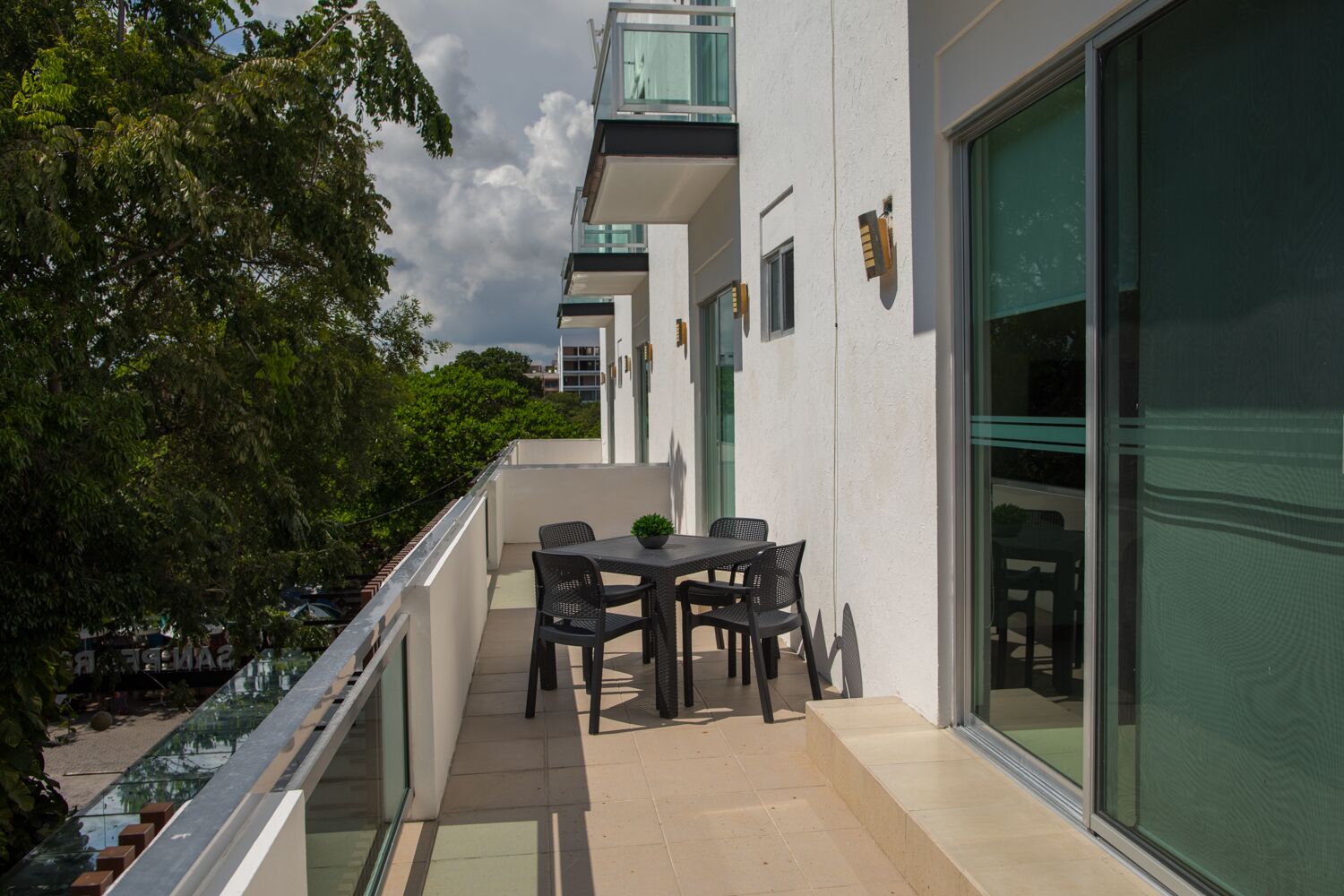 Un balcon avec une table et des chaises, une rambarde en verre et une vue sur les arbres et les bâtiments.