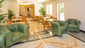 Un hall d’hôtel doté de canapés en cuir vert, de tables et de chaises en bois, d’un sol en marbre et de grandes fenêtres laissant entrer la lumière naturelle.